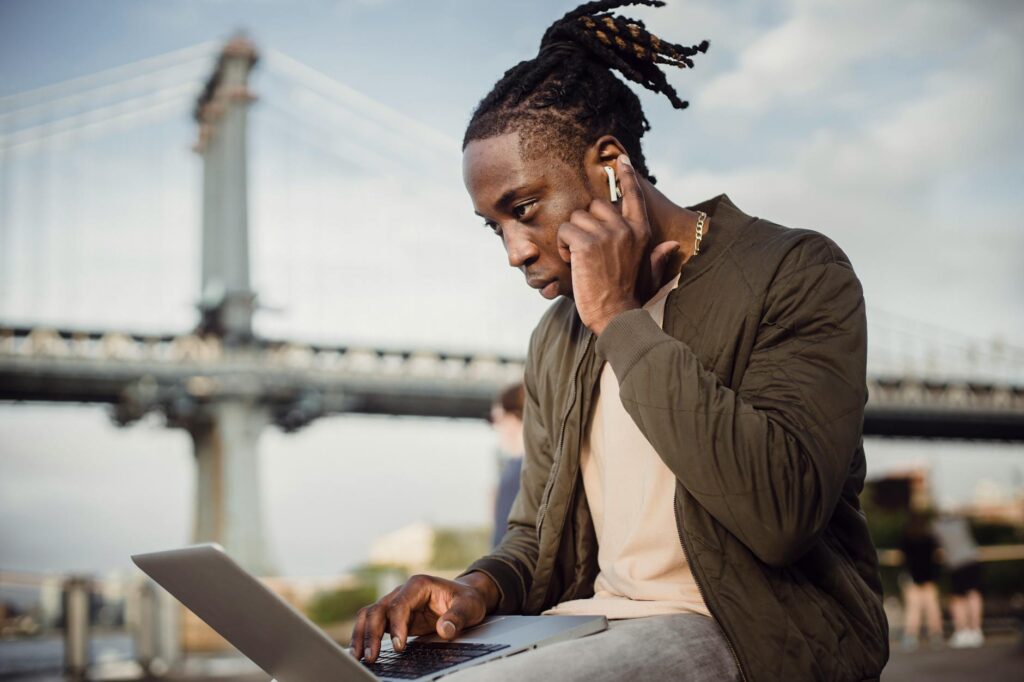 Low angle of concentrated black man in casual wear using laptop and earphones while working outdoors in park