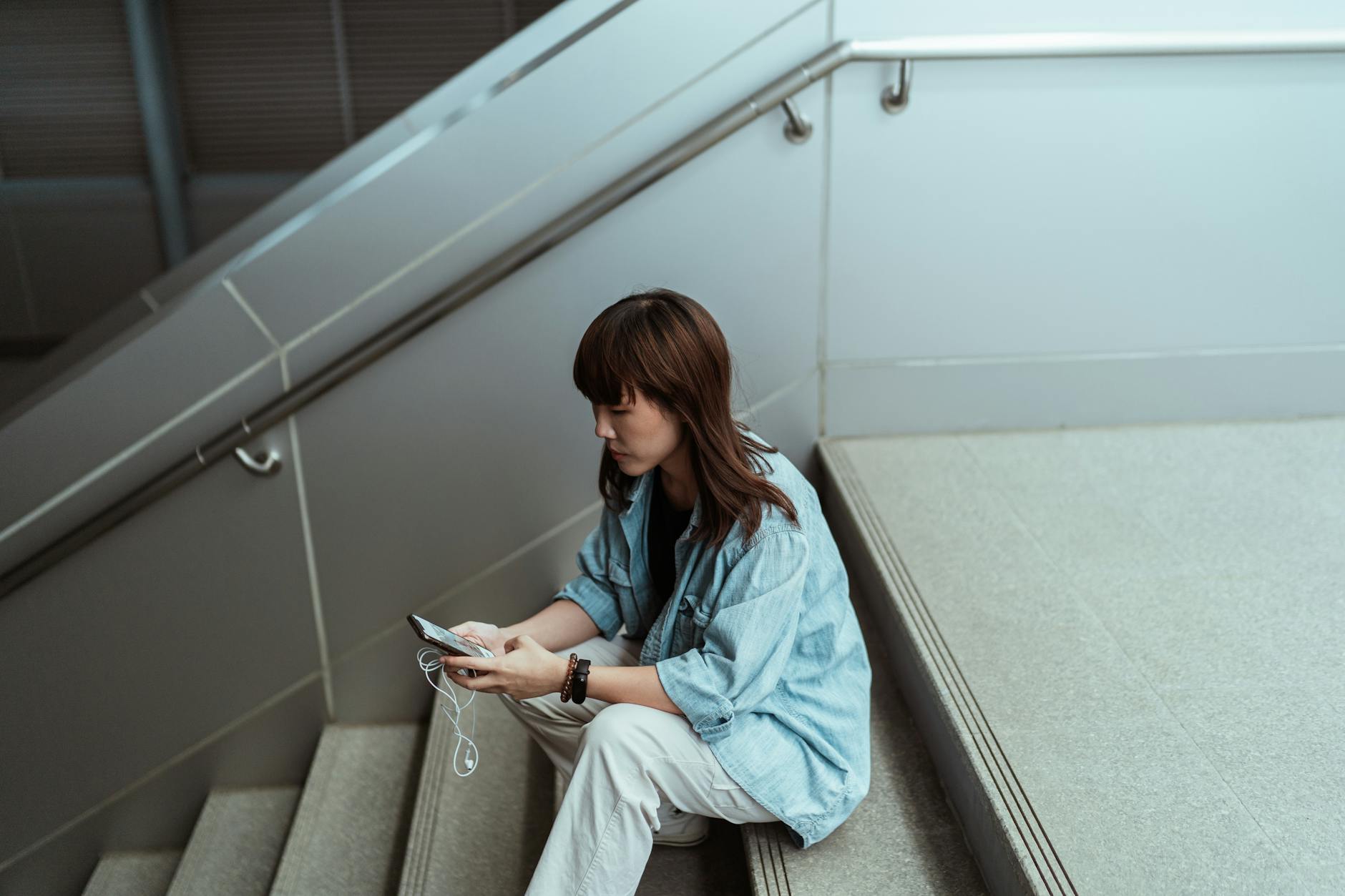 From above side view of pensive Asian female in casual clothes browsing internet on cellphone while sitting on stairs with earphones in building