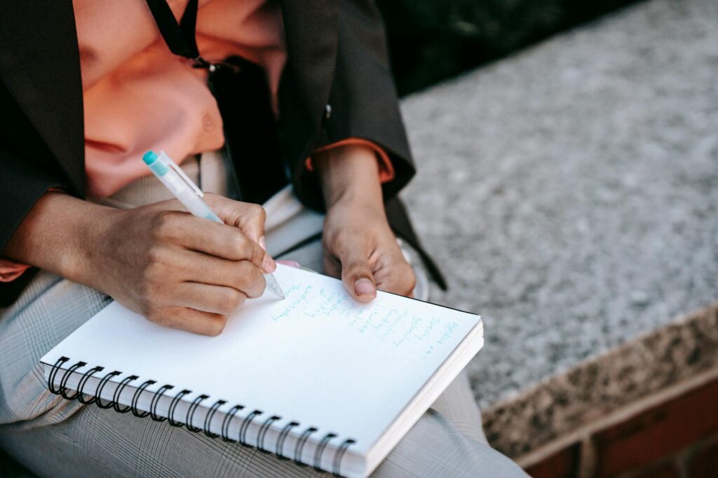 From above of crop anonymous ethnic woman making list in notebook during work in park