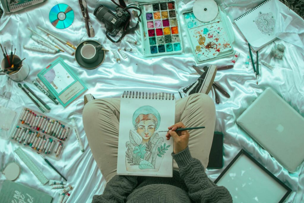 Person Painting a Female Portrait While Sitting on a Floor with Various Arts Items around