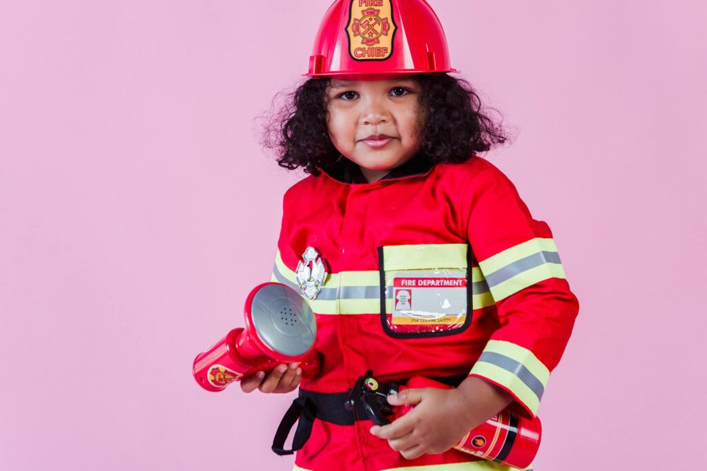 Little ethnic child wearing fireman costume with fire extinguisher and loudspeaker toy with hardhat and standing on pink background and looking at camera